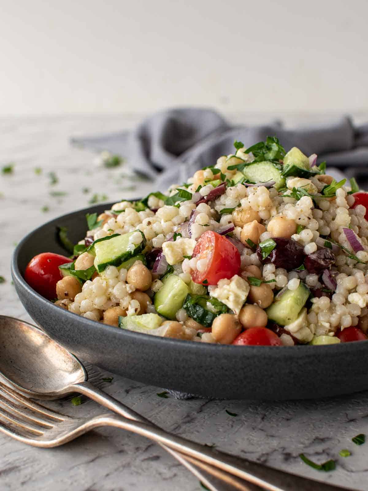 bowl of Greek couscous salad with fork and spoon and grey towel in the background.