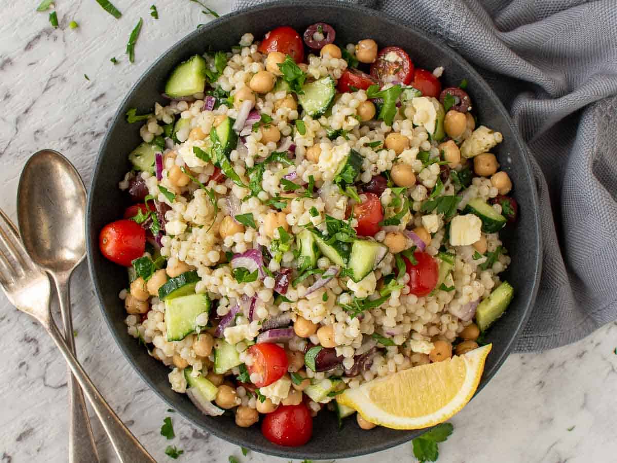 A bowl of couscous salad with fork and spoon on the side and grey towel viewed from above.