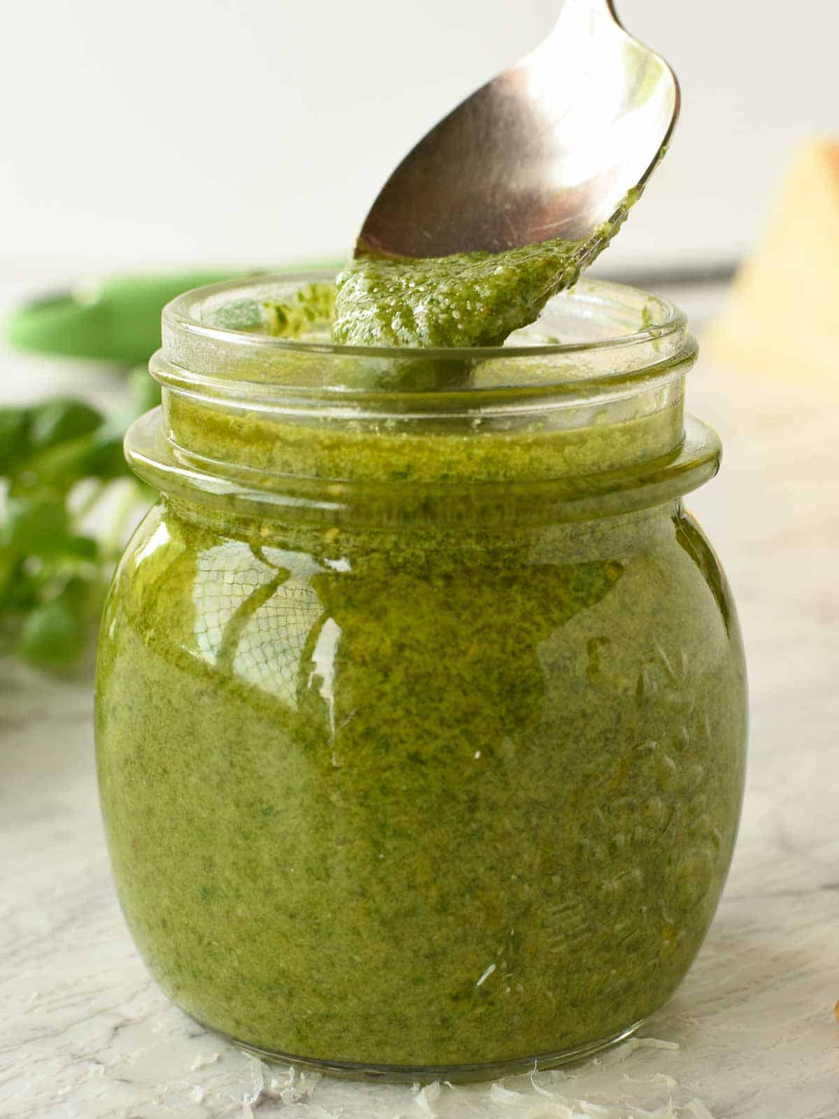 glass jar of pesto with a spoonful being spooned out, fresh basil in the background