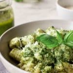 image with text. text reads "homemade pesto gnocchi". a bowl of pesto gnocchi on a grey cloth with jar of pesto in the background.