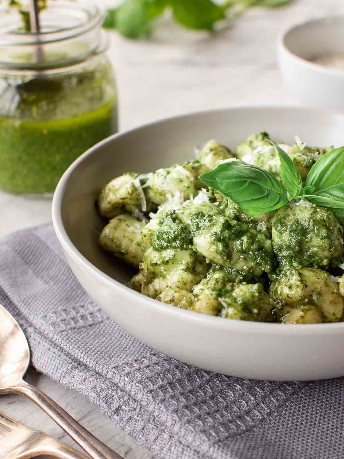 A bowl of pesto gnocchi on a grey cloth with jar of pesto in the background.