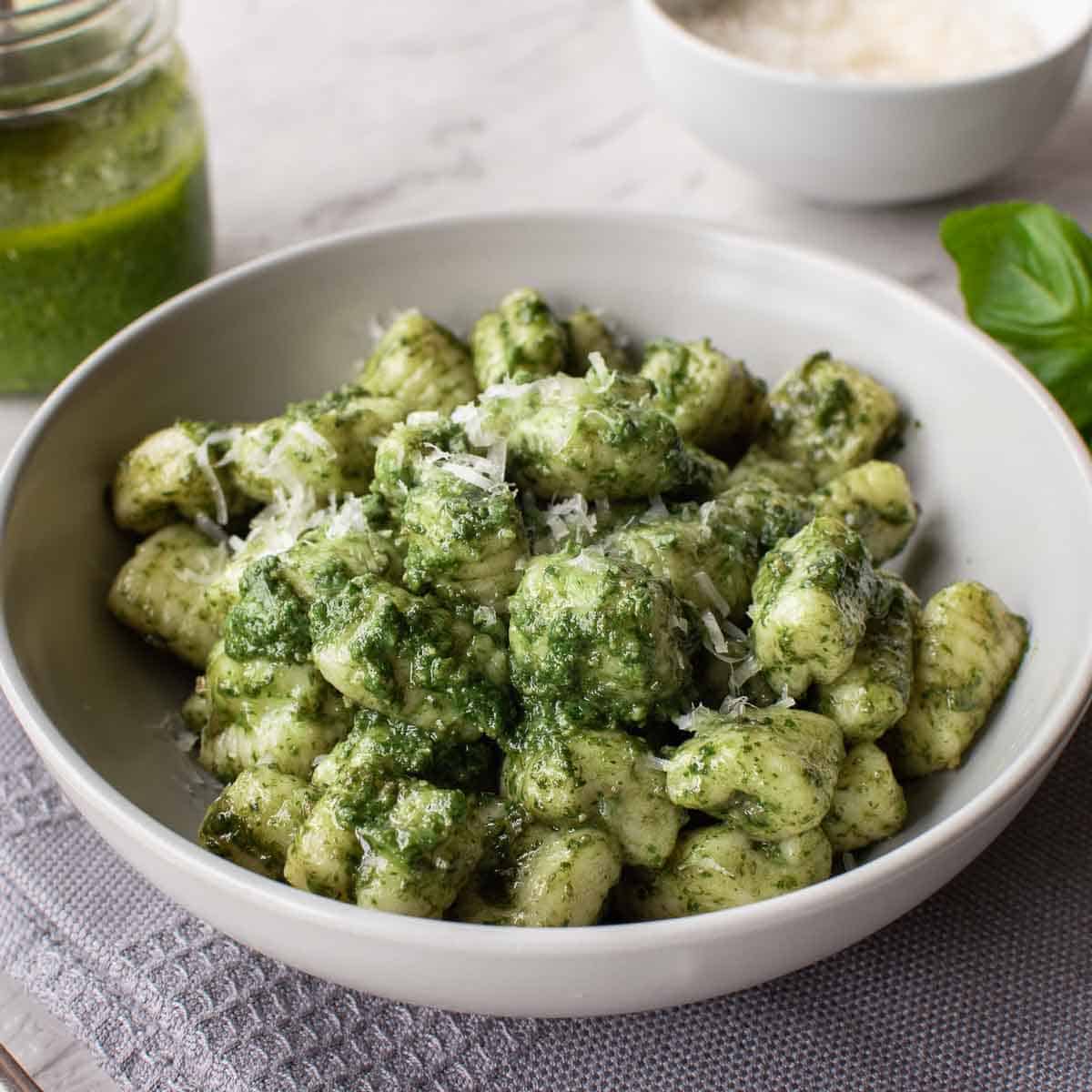 a bowl of pesto gnocchi on a grey cloth with jar of pesto and bowl of parmesan in the background.
