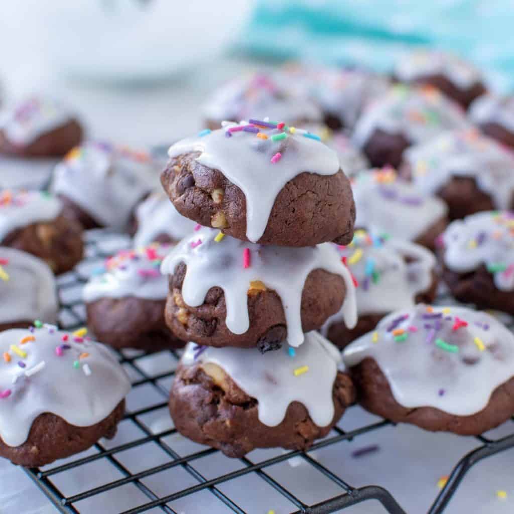close up of three chocolate cookies with white frosting and sprinkles stacked with extra cookies around all on a wire rack.