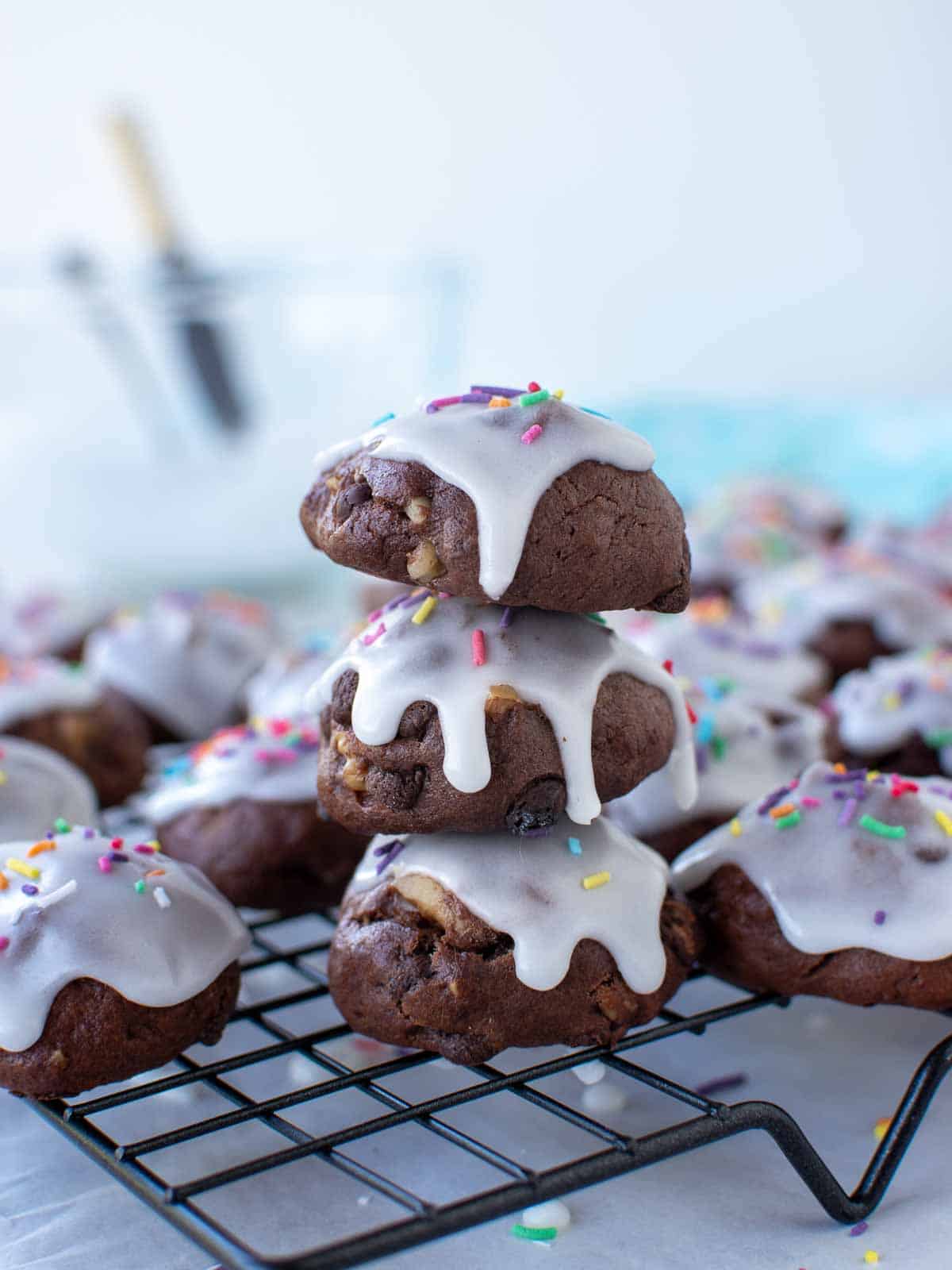 three chocolate cookies with white frosting and sprinkles stacked with extra cookies around all on a wire rack.