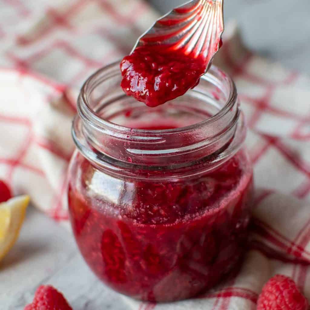 raspberry sauce in a glass jar with a spoon full lifted out.