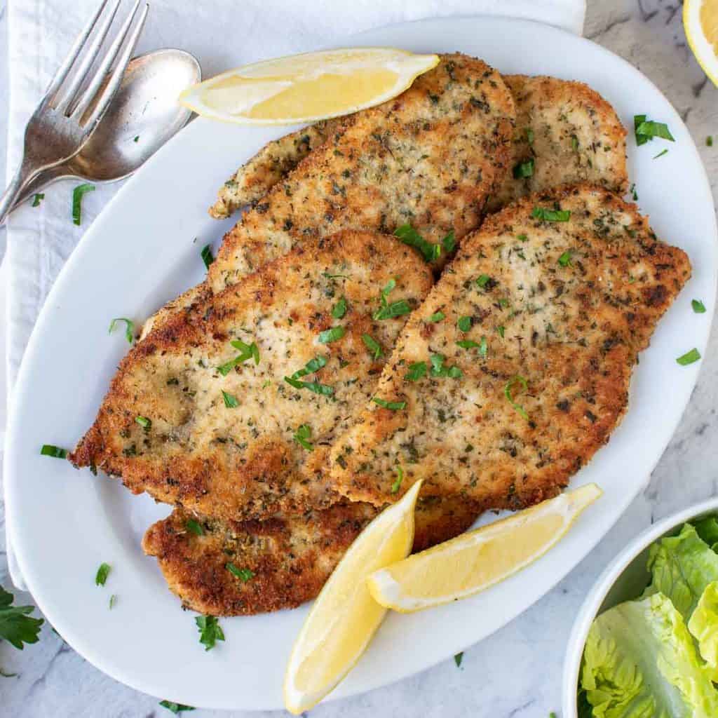 browned chicken cutlets on white plate with lemon wedges viewed from above.