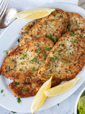 browned chicken cutlets on white plate with lemon wedges viewed from above.