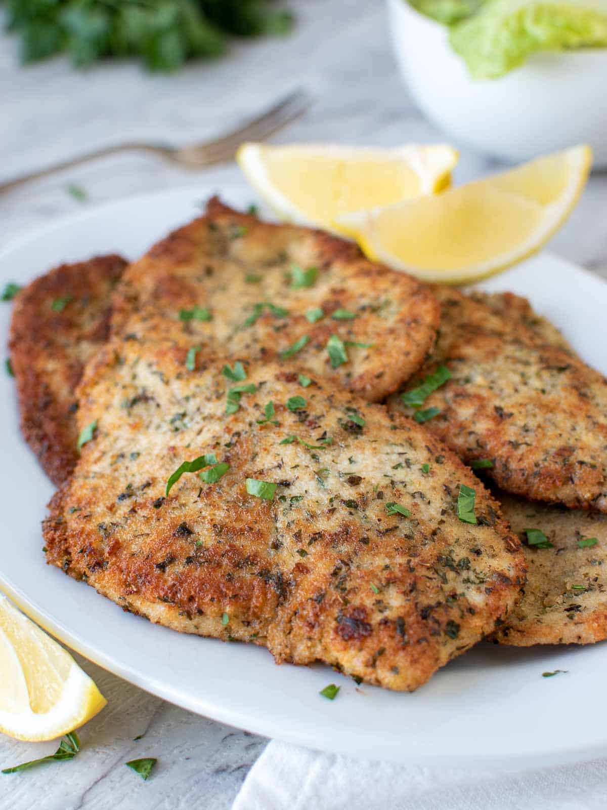 Italian chicken cutlets on white plate with lemon wedges and chopped parsley garnish.