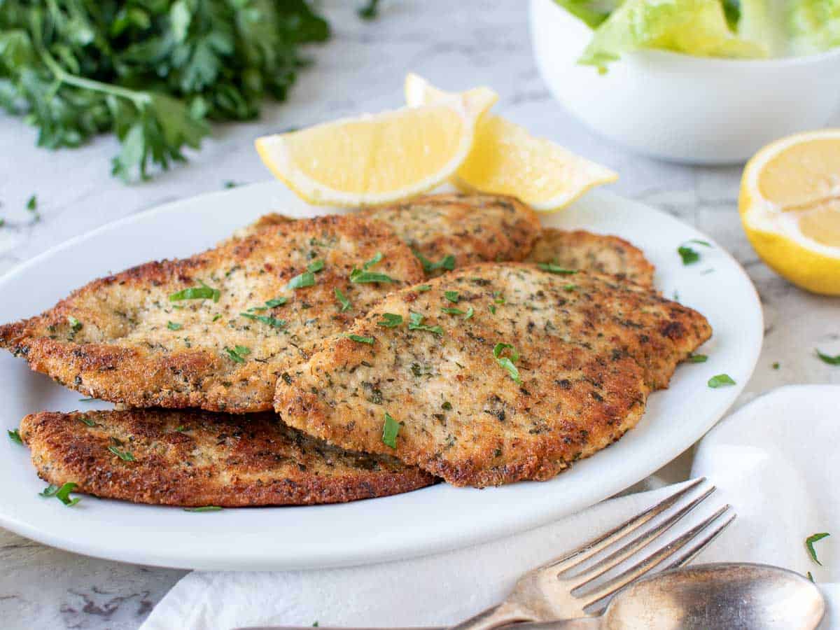 Browned chicken cutlets on white plate with lemon wedges, parsley and salad in the background.