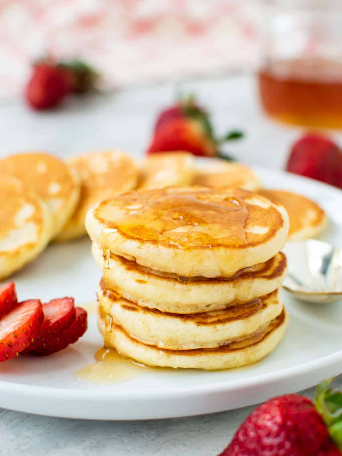 Four mini pancakes stacked on a white plate with syrup dripping off and more pancakes and strawberries around.