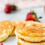 image with text. text reads "fast and easy mini pancakes". image is four mini pancakes stacked on a white plate with syrup dripping off and more pancakes and strawberries around.