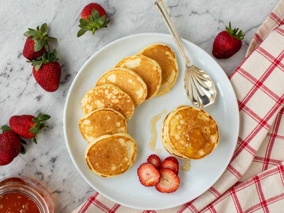 Four pancakes stacked on a white plate with syrup dripping off and more pancakes and strawberries around viewed from above.