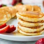 four mini pancakes stacked on a white plate with syrup dripping off and more pancakes and strawberries around.