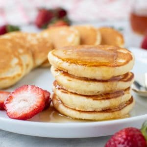 close up of four mini pancakes stacked on a white plate with syrup dripping off and more pancakes and strawberries around.