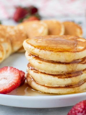 close up of four mini pancakes stacked on a white plate with syrup dripping off and more pancakes and strawberries around.