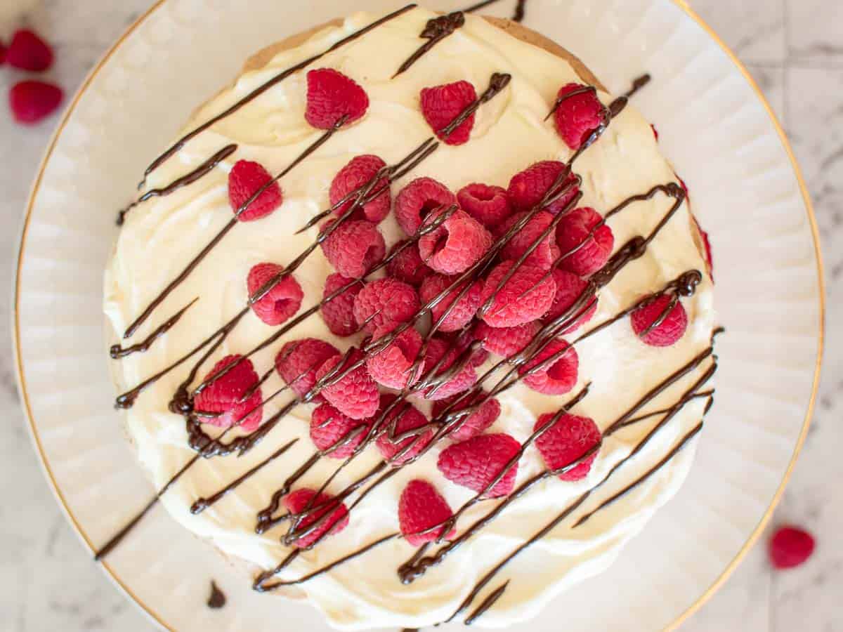 a cream and raspberry cake drizzled with chocolate viewed from above on a white plate.