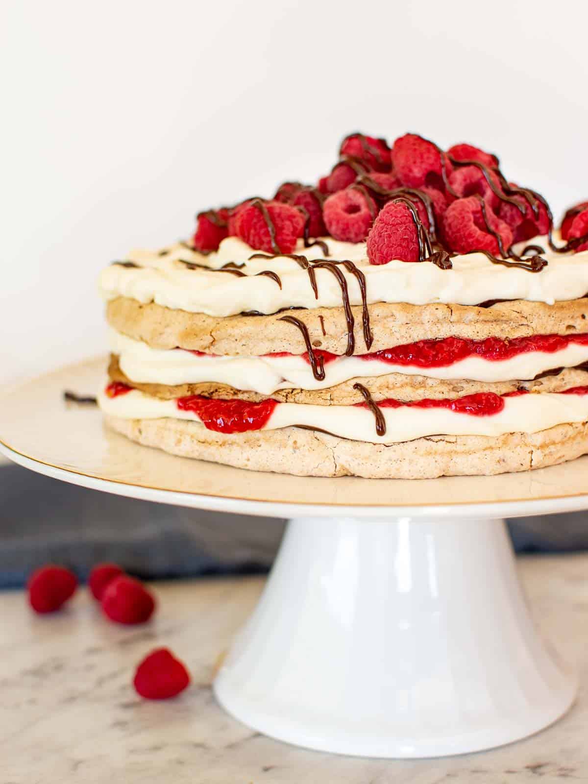three layer meringue cake with cream, raspberries and chocolate on a white cake stand with raspberries scattered underneath.