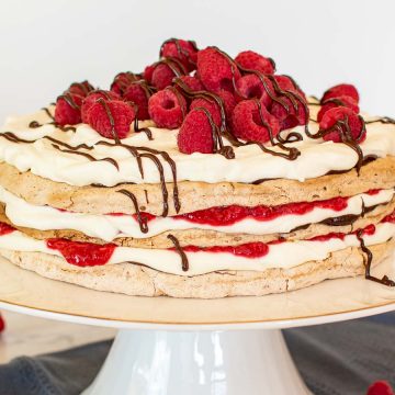 close up of three layer meringue cake with cream, raspberries and chocolate on a white cake stand with raspberries scattered underneath.