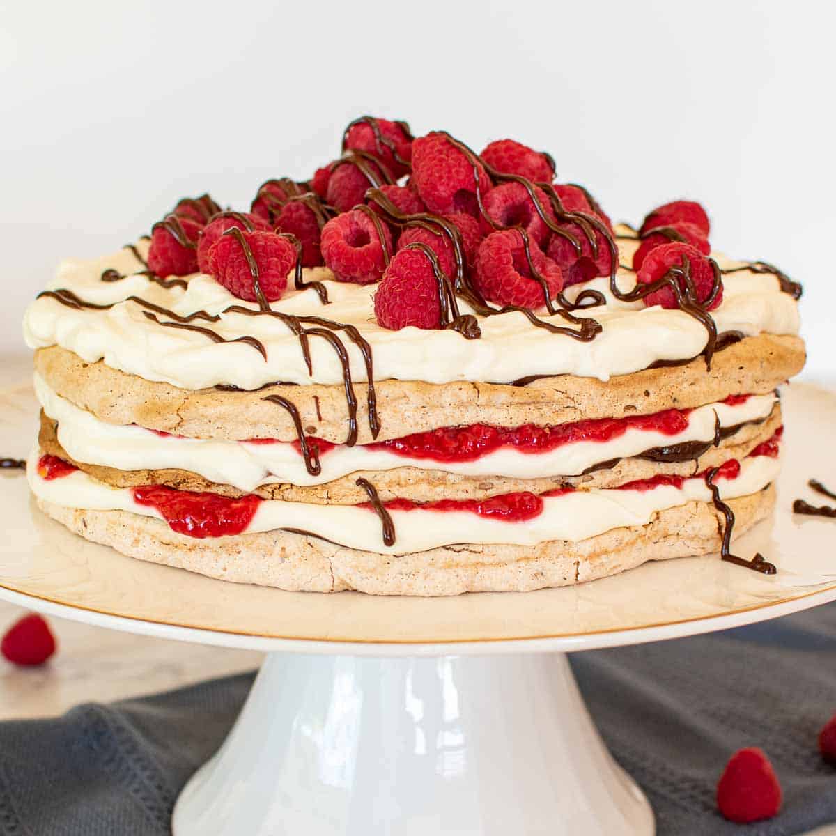 three layer meringue cake with cream, raspberries and chocolate on a white cake stand with raspberries scattered underneath.