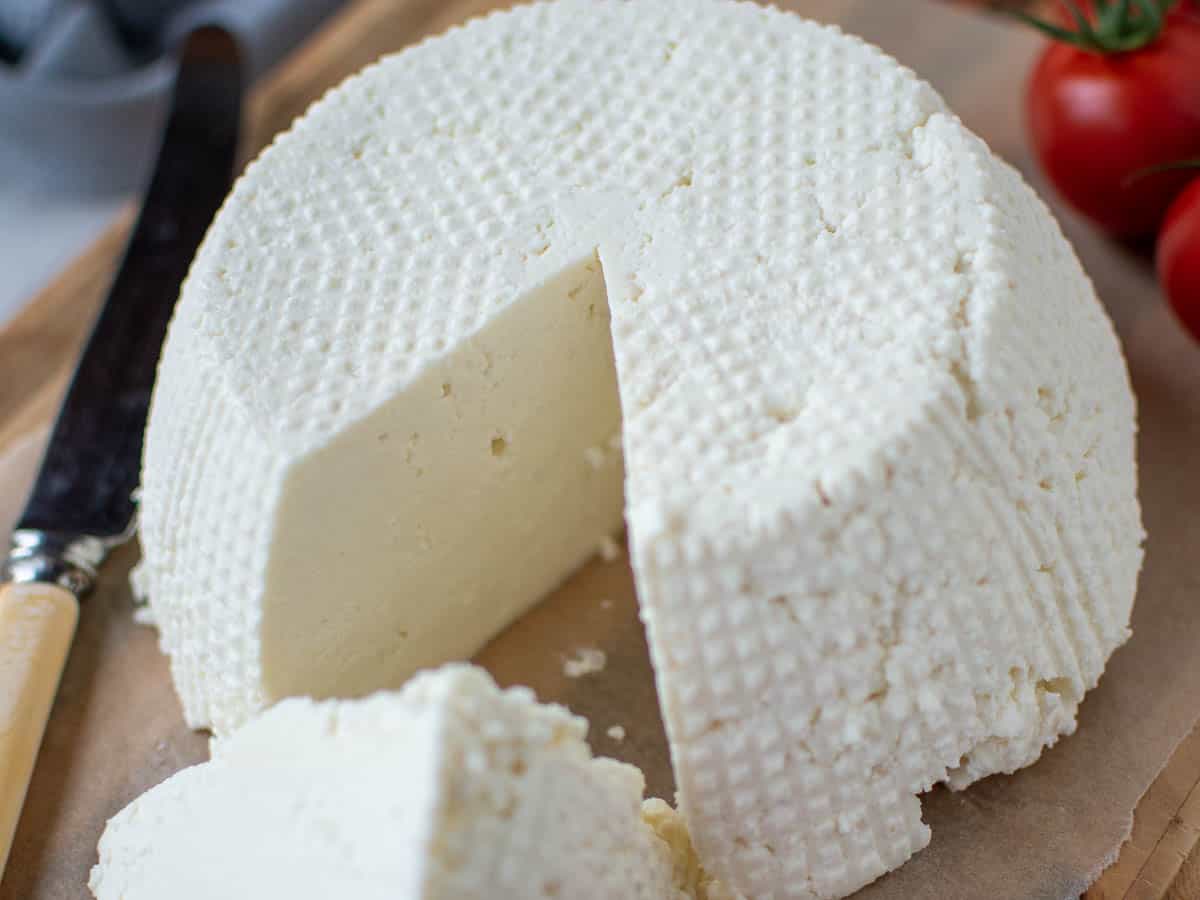 whole fresh ricotta cheese on wooden board with a wedge cut out; wedge in front of whole cheese.