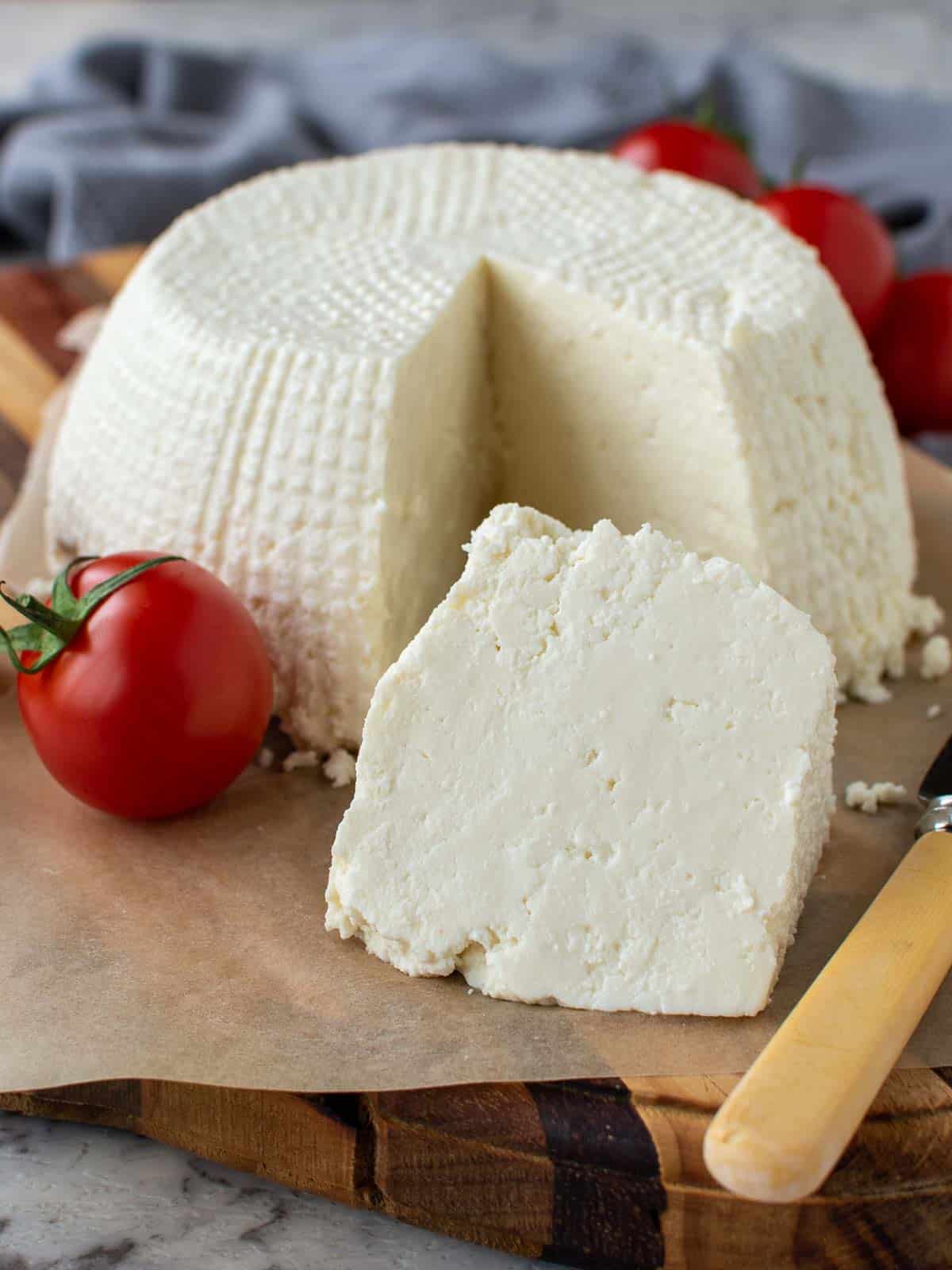 whole fresh ricotta cheese on wooden board with a wedge cut out; wedge in front of whole cheese.