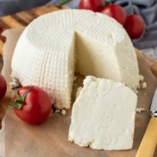 whole fresh ricotta cheese on wooden board with a wedge cut out; wedge in front of whole cheese.