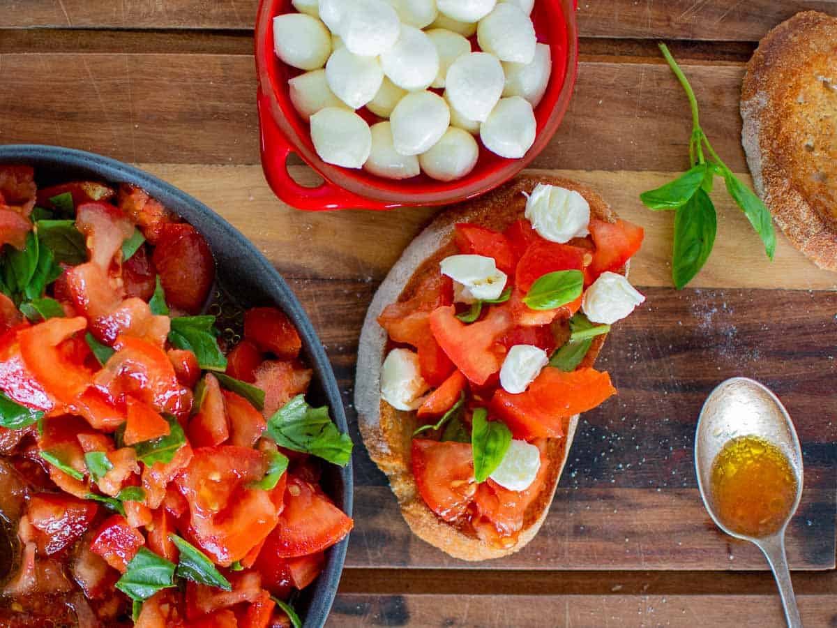 Bruschetta with tomatoes and mozzarella, red bowl filled with bocconcini, dark grey bowl with chopped tomatoes and basil, spoon and basil sprig all on wooden board viewed from above.