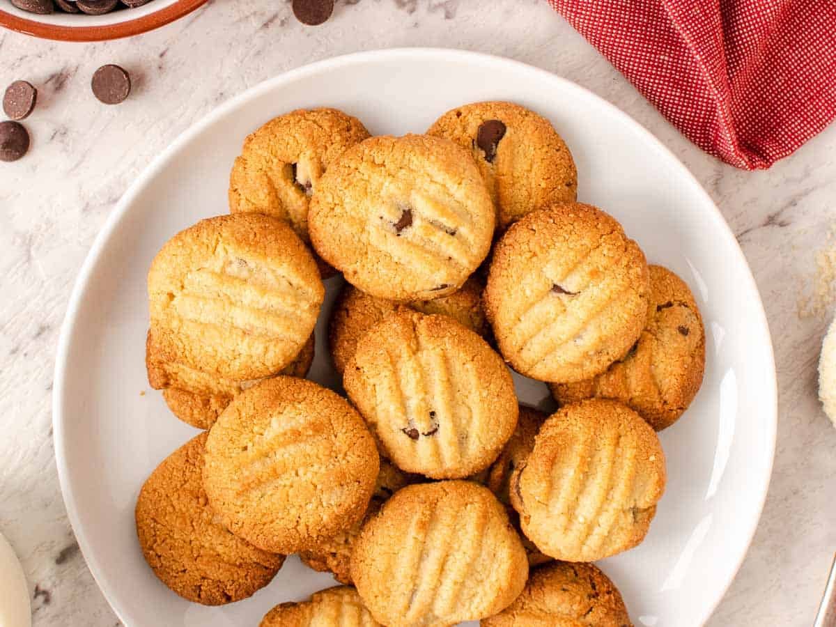 The cookies on a white plate with chocolate chips and almonds scattered nearby viewed from above.