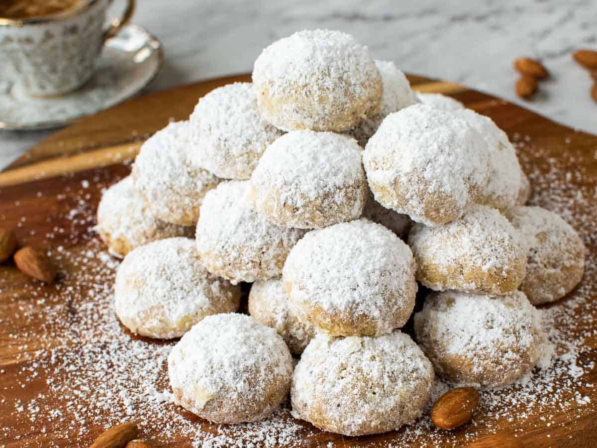 Pile of white sugar coated cookies on wooden board with espresso cup in the background.