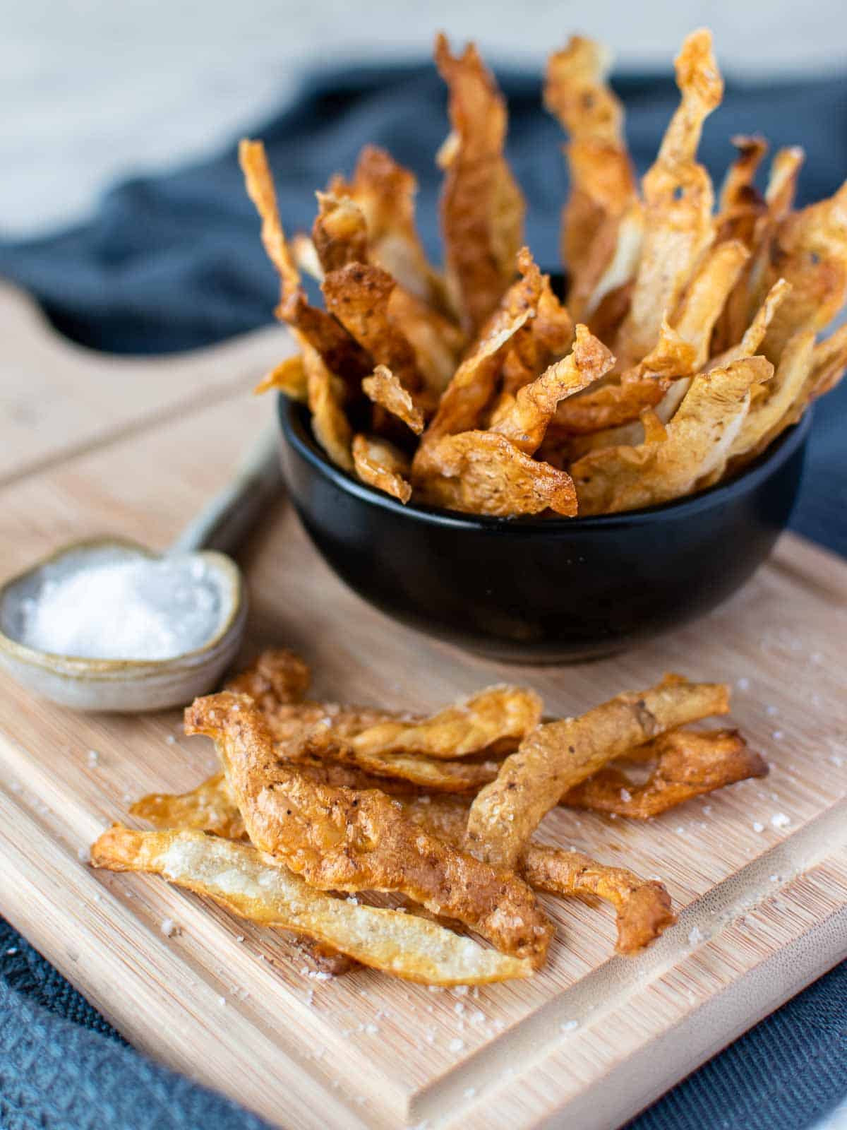 potato skin chips piled into black bowl placed onto a wooden board with more chips on the board and a spoon filled with salt.