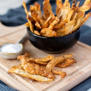 potato skin chips piled into black bowl placed onto a wooden board with more chips on the board and a spoon filled with salt.