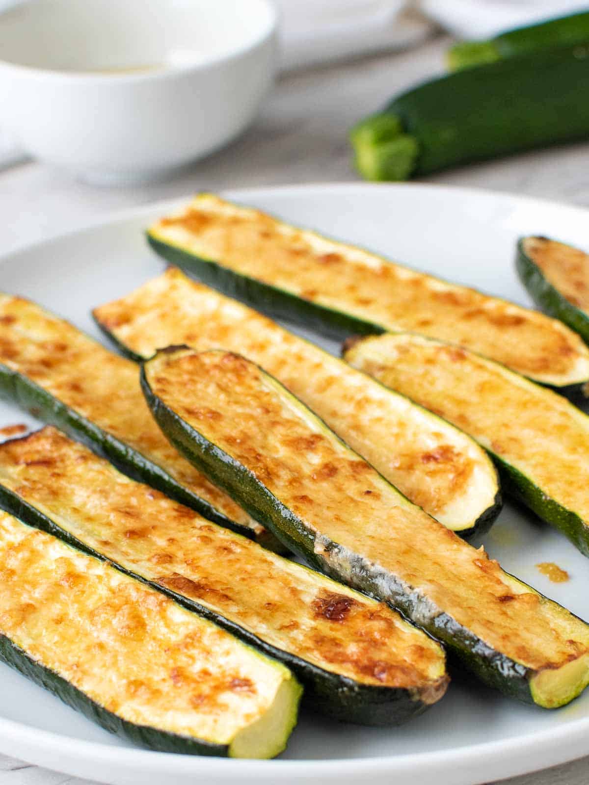 Pile of roasted zucchini on a white plate with fresh zucchini in background.