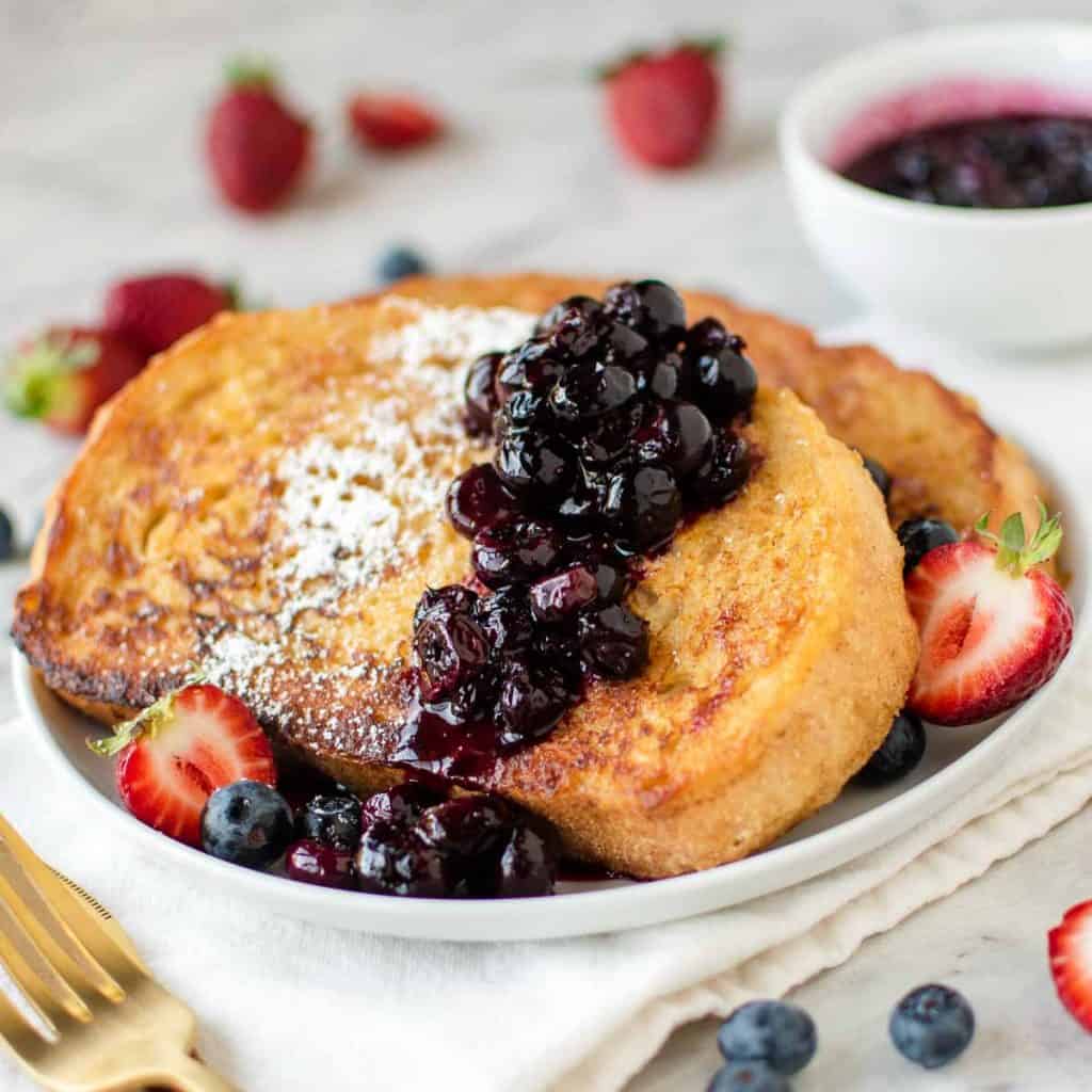 Two slices of sourdough French toast with blueberry compote, powdered sugar, and halved strawberries on a white plate set on a table next to cutlery.