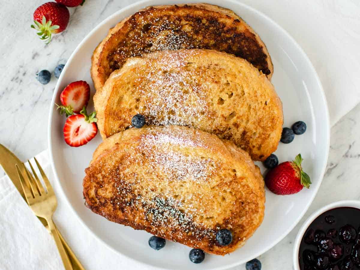 Three slices of the finished french toast recipe on a white plate with strawberries and blueberries.