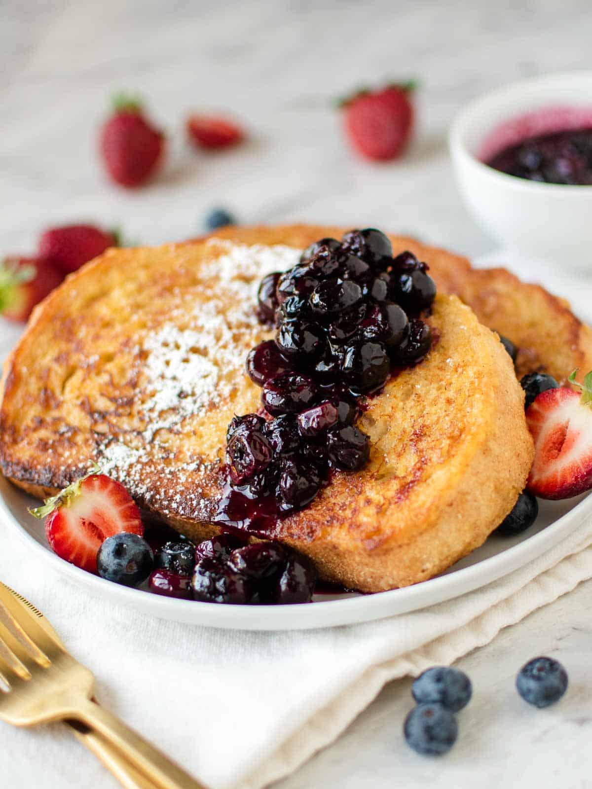 Two slices of sourdough French toast with blueberry compote, powdered sugar, and halved strawberries on a white plate set on a table next to cutlery.