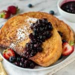 Two slices of sourdough French toast with blueberry compote, powdered sugar, and halved strawberries on a white plate set on a table next to cutlery.