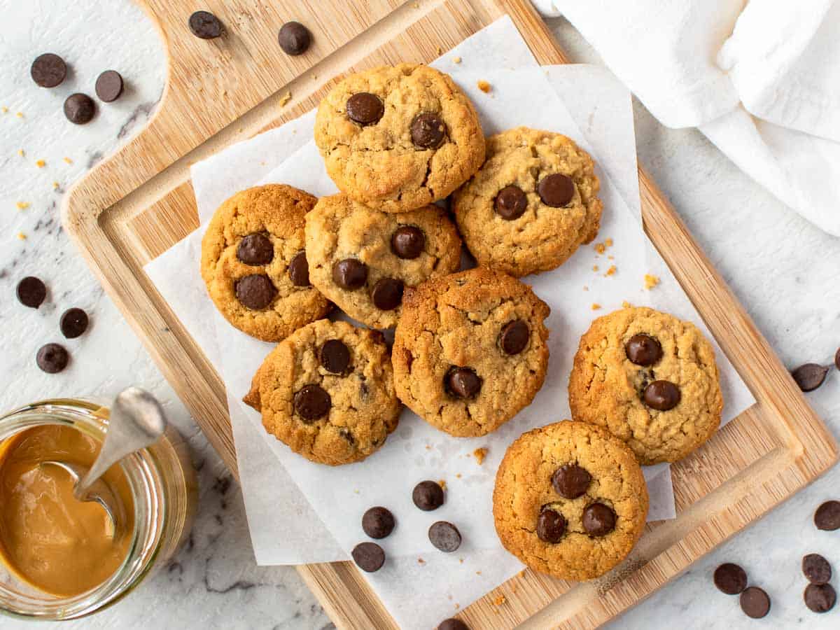 Cookies with chocolate chips on a wooden board surrounded by chocolate chips and a glass jar of peanut butter.