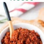 Text and image. text reads sun dried tomato pesto rosso. image is sun dried tomato pesto in a white bowl with small wooden spoon in the pesto.