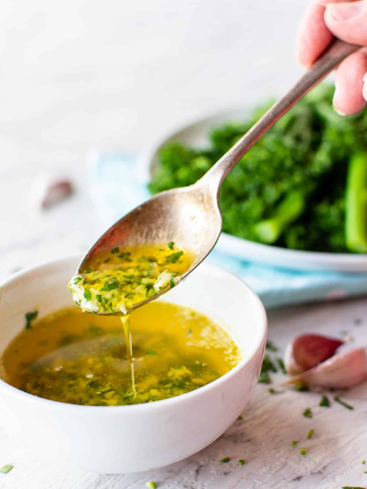 A spoonful of garlic butter sauce taken from a small white bowl with garlic and broccolini in the background.