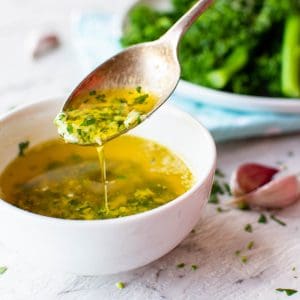 A silver teaspoonful of garlic butter sauce taken from a small white bowl with garlic and broccolini in the background.