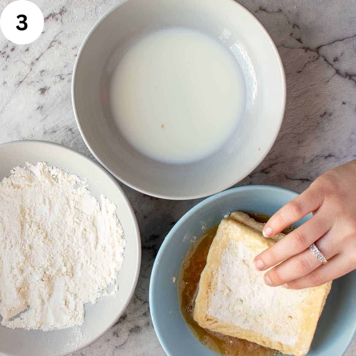 Dipping a sandwich in beaten egg with bowl of milk and bowl of flour on the side.