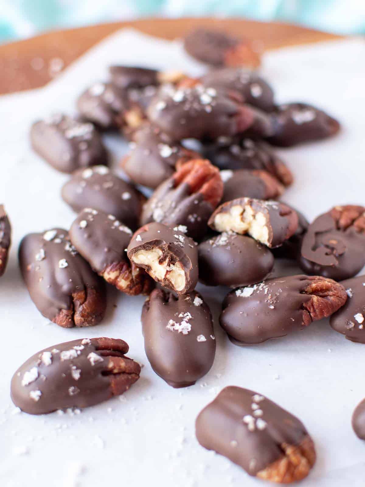 Pile of chocolate dipped pecans sprinkled with flaked salt and resting on white parchment paper.