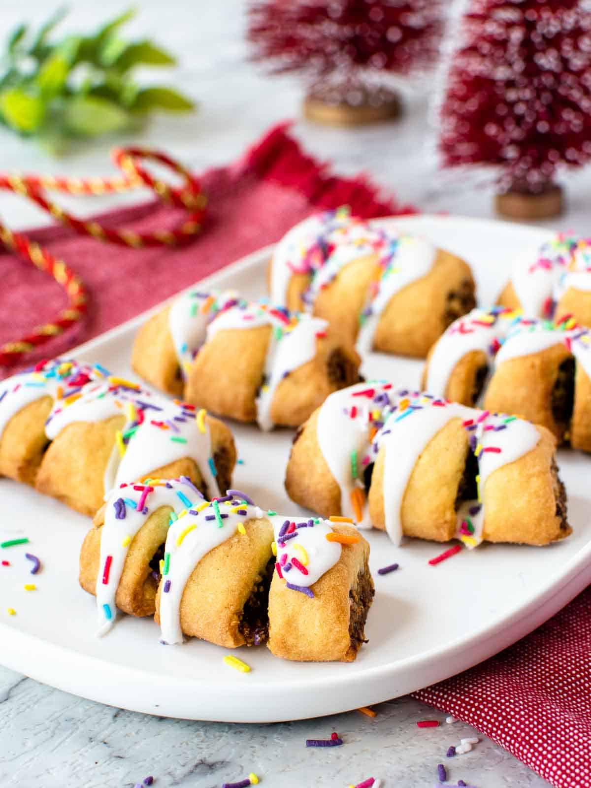 seven cuccidati cookies with a dark fruit filling drizzled with white icing and colored sprinkles on a white oblong plate.