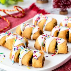 seven cuccidati cookies with a dark fruit filling drizzled with white icing and colored sprinkles on a white oblong plate.