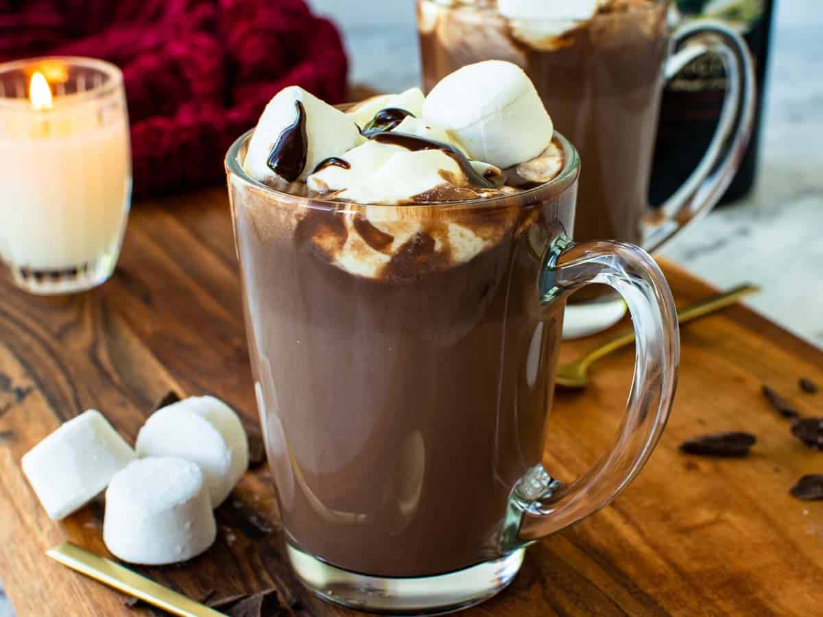 A clear glass mug filled with hot chocolate topped with marshmallows and cream. A second mug sits behind the first.