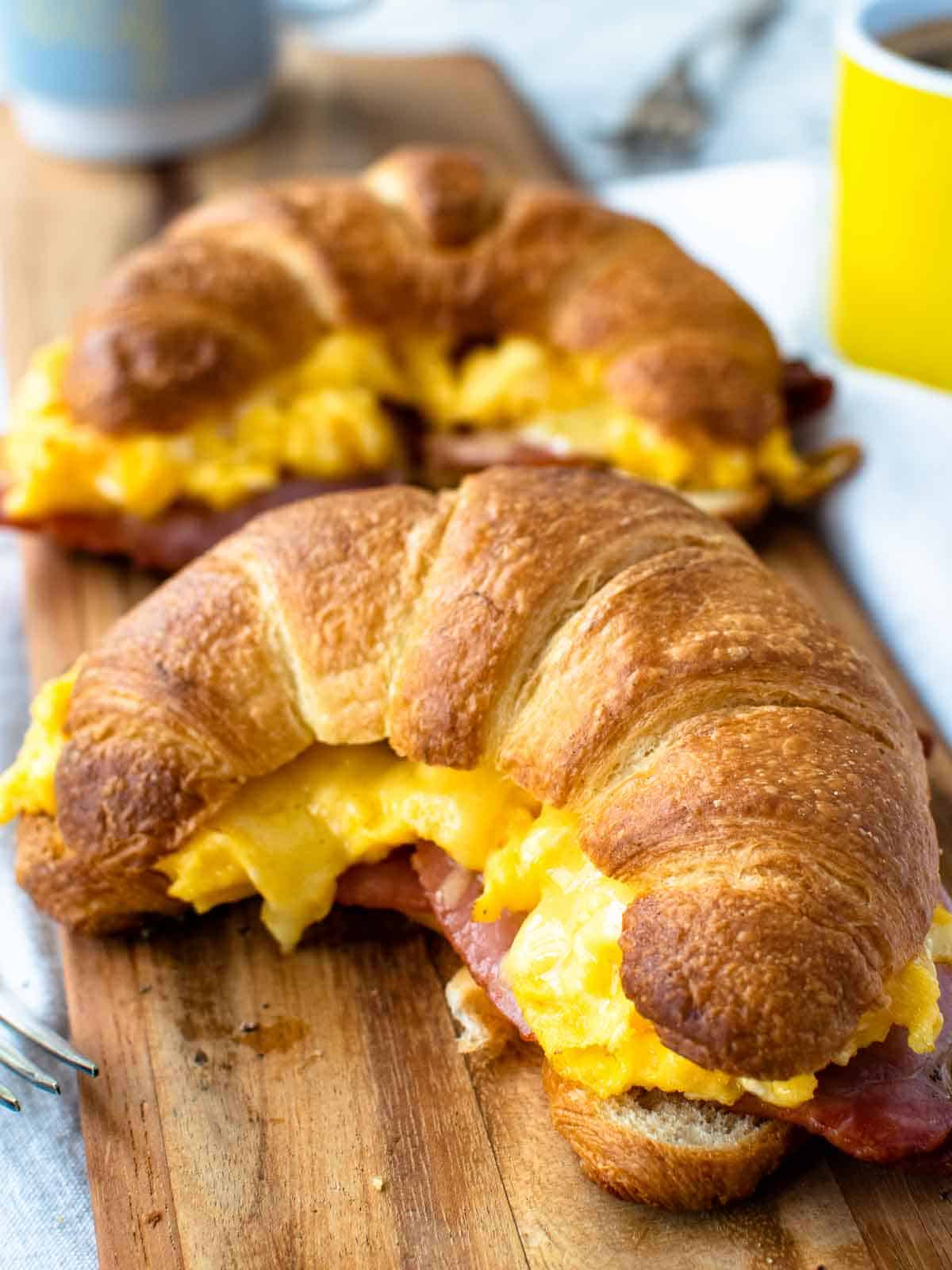 Two bacon egg and cheese croissants on a wooden board with cups in the background.