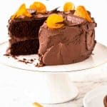 text reads "delicious layered chocolate orange cake". image is a two layer chocolate cake on white stand with slice of cake on white plate.