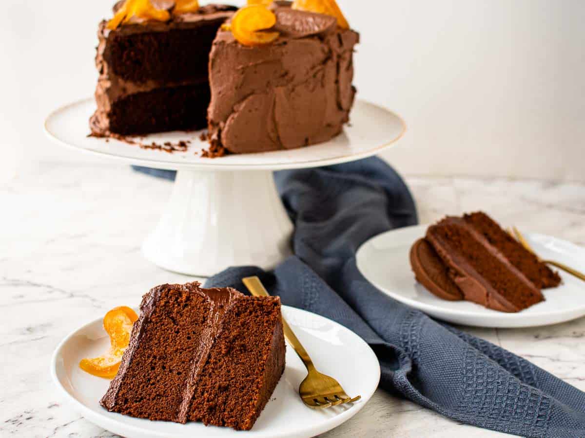 A two layer chocolate cake on white stand with two slices on two white plates.