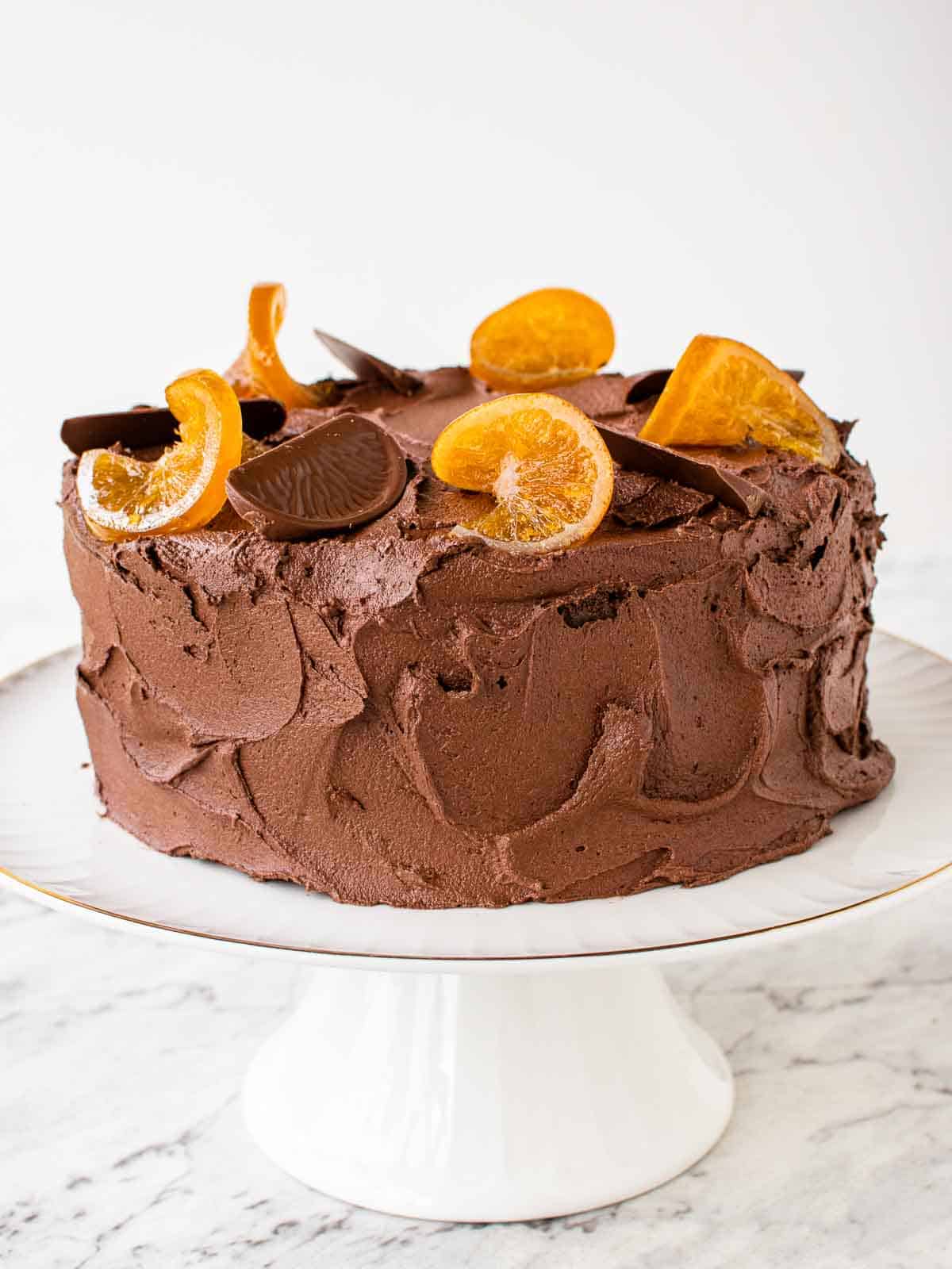 Chocolate orange cake on white cake stand with orange slices decoratively placed on top.