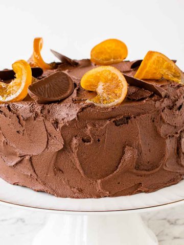 brown frosted cake on white cake stand with orange slices decoratively placed on top.
